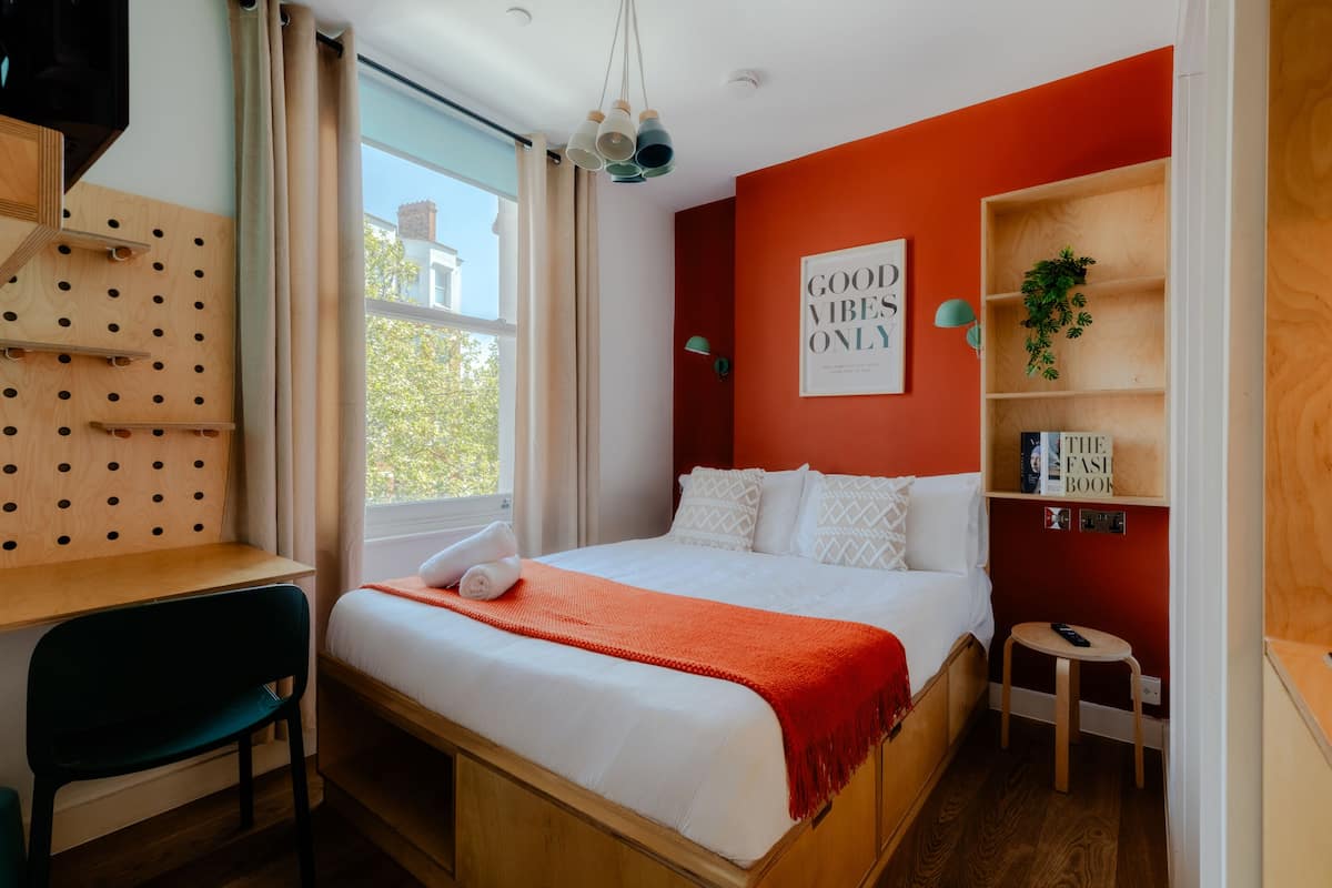 A bedroom with a bed, a desk, a chair, a small stool, a shelf with books, and a window with curtains.