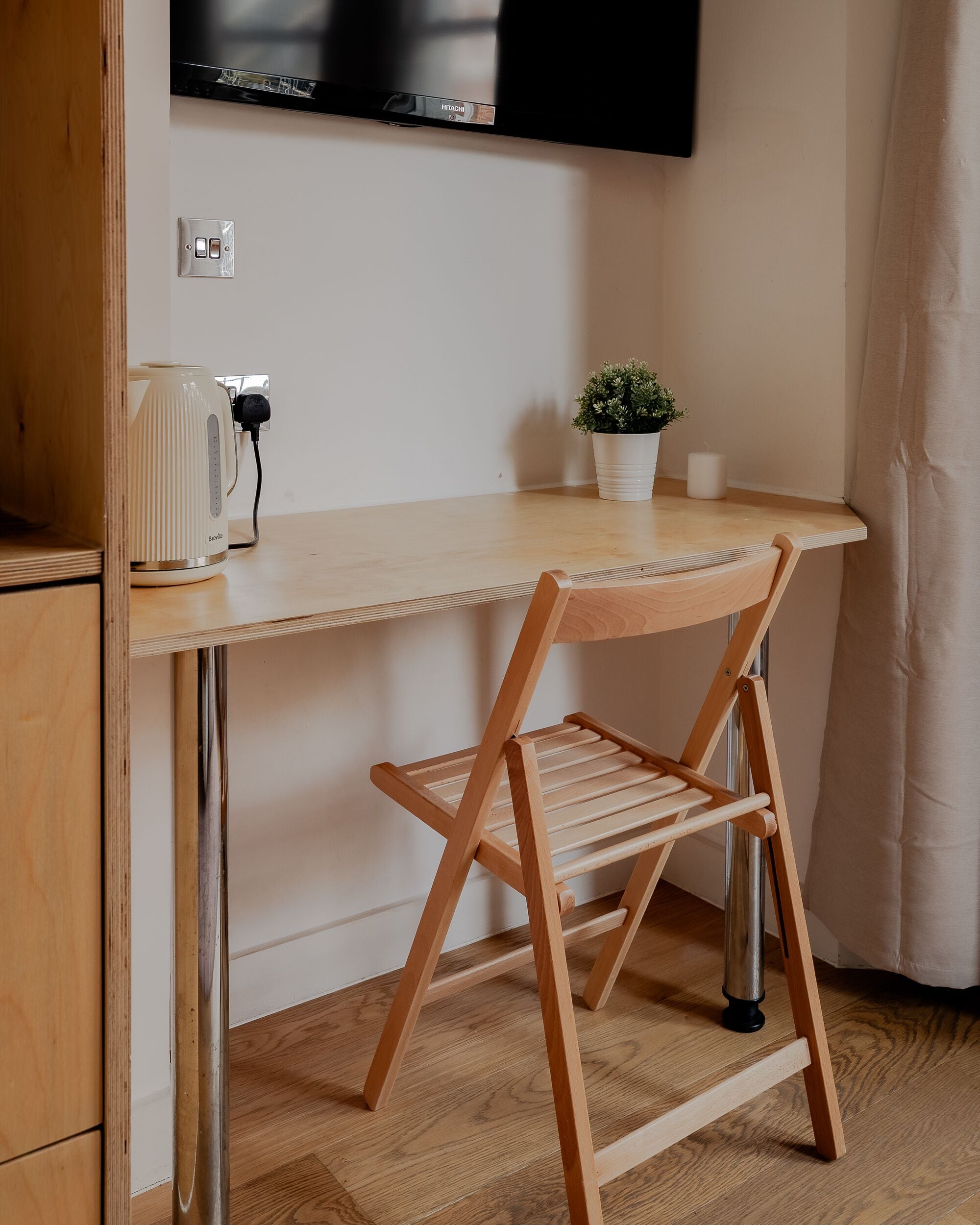 A compact workspace with a wooden desk, a Breville coffee maker, a potted plant, and a foldable chair.