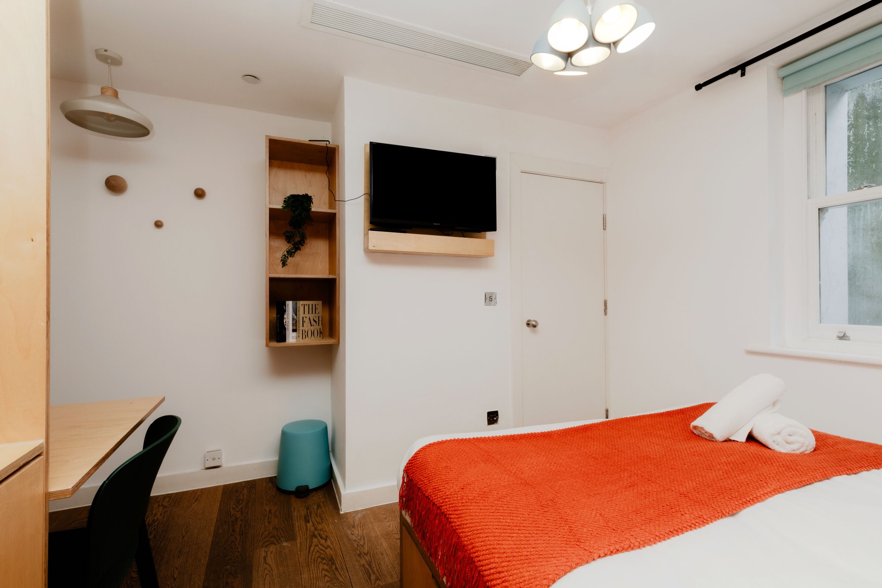 A bedroom with a bed, a desk, a chair, a television, and a window.