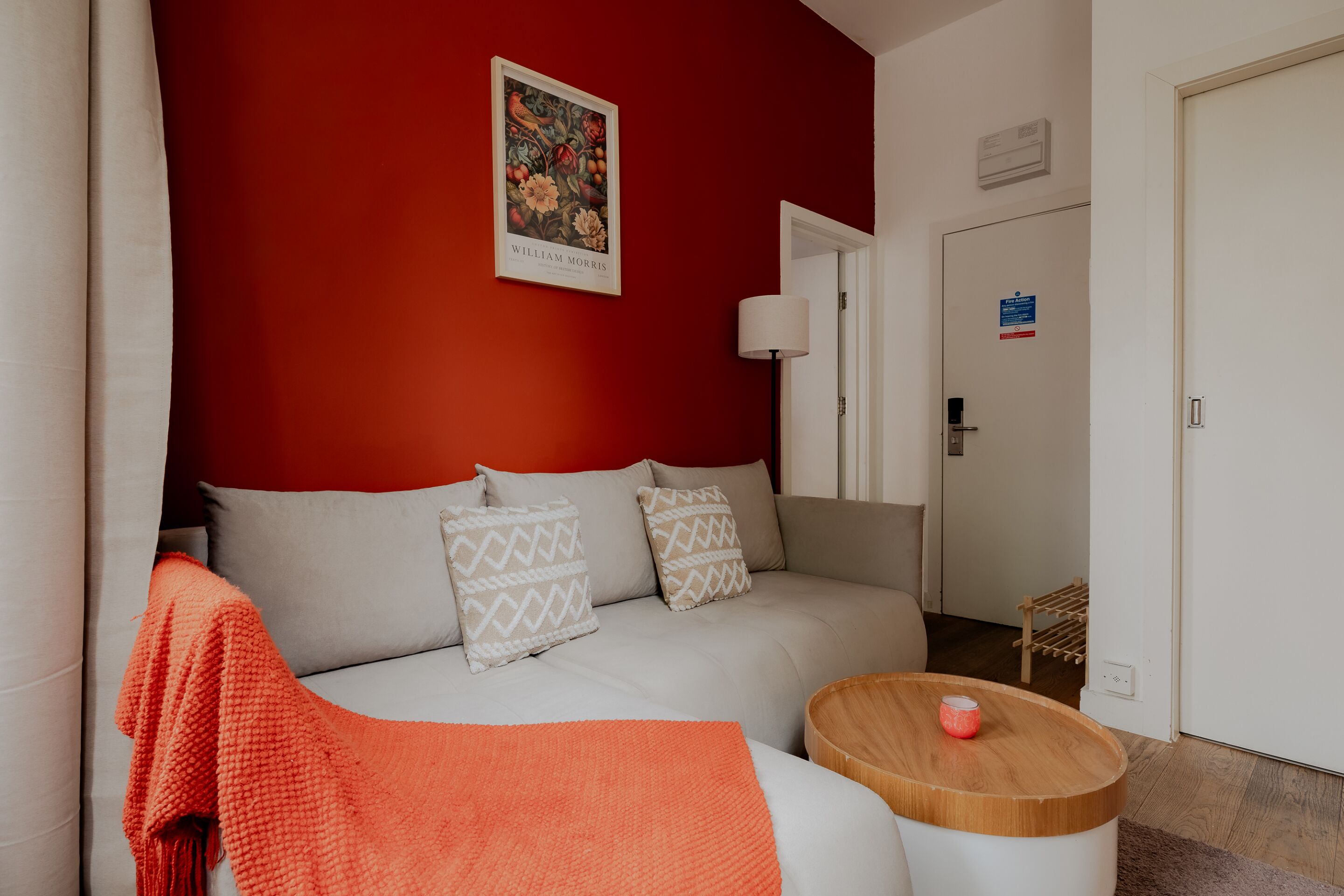 Un salon moderne avec un mur d’accent rouge, un canapé blanc agrémenté de coussins à motifs, une table basse en bois et une porte portant un panneau indiquant les consignes de sécurité en cas d’incendie.