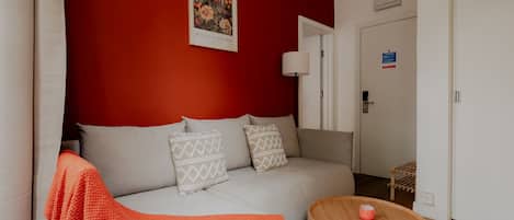 Un salon moderne avec un mur d’accent rouge, un canapé blanc agrémenté de coussins à motifs, une table basse en bois et une porte portant un panneau indiquant les consignes de sécurité en cas d’incendie.