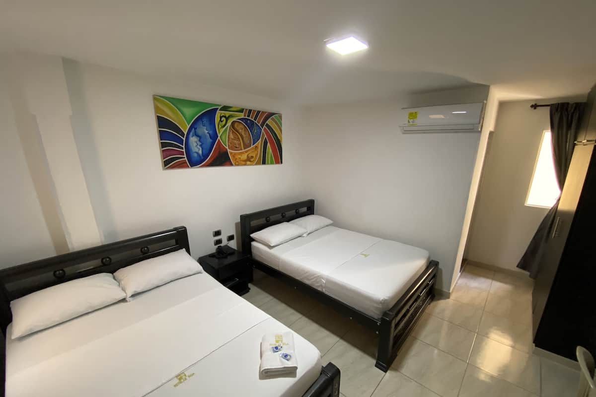 Une chambre d’hôtel avec deux lits simples, une table de chevet, un climatiseur fixé au mur et un tableau abstrait aux couleurs vives.