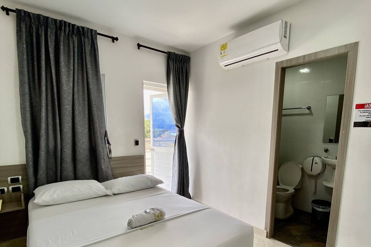 Une chambre d’hôtel avec un lit, une salle de bain visible à travers une porte ouverte et un appareil de climatisation fixé au mur.