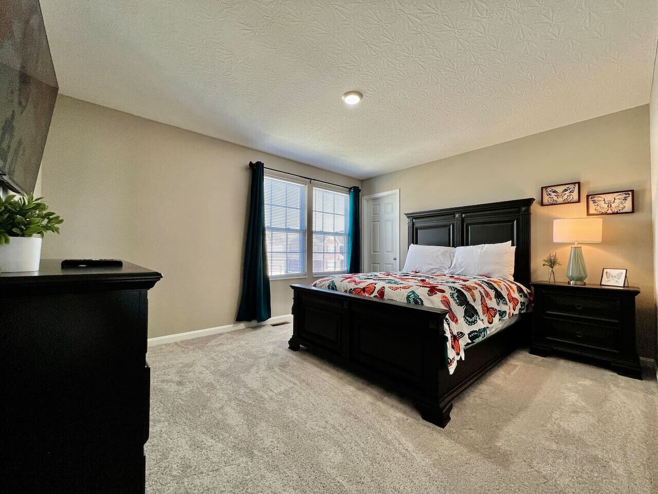 A bedroom with a bed, a dresser, a nightstand, a lamp, a window with curtains, and a plant.