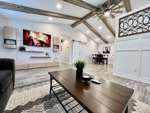A modern living room with a wooden dining table, a flat-screen TV, and a ceiling fan.