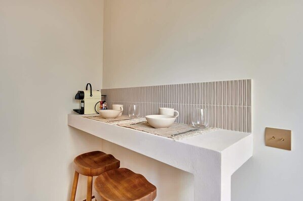 Une cuisine avec un plan de travail blanc, deux tabourets en bois et un mur recouvert de carreaux.