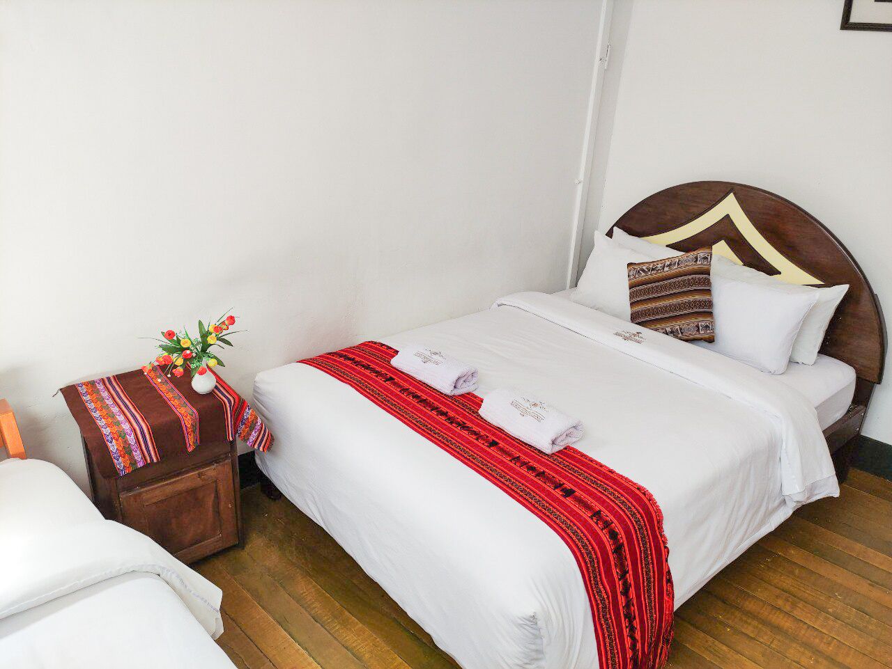 A neatly made bed with white linens and a red and black patterned blanket, a wooden nightstand with a vase of flowers, and a wooden headboard with a decorative pillow.