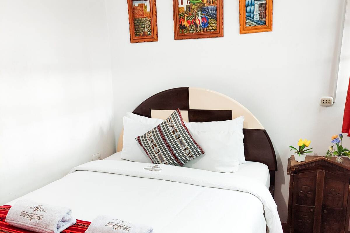 A neatly made bed with white linens and a red patterned throw, a wooden headboard, and three framed paintings on the wall.