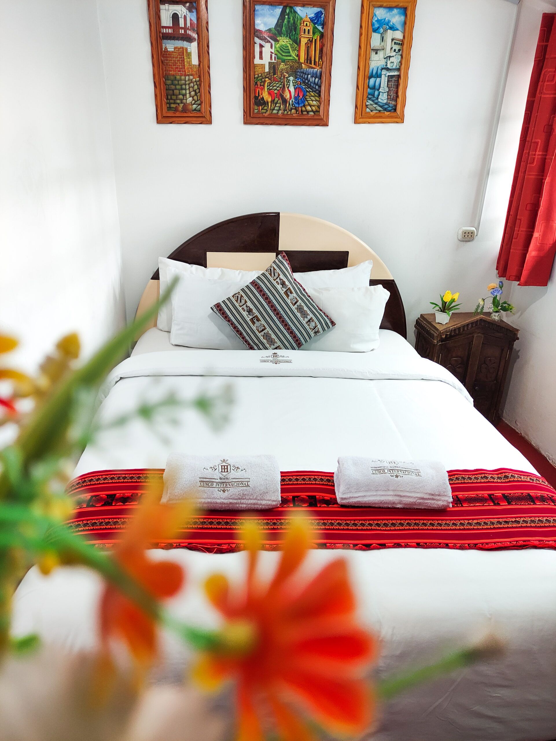 A neatly made bed with white linens, a patterned pillow, and two folded towels with a logo. There are three framed paintings on the wall above the bed.