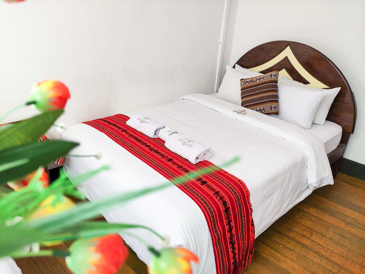 A neatly made bed with white linens and a red patterned throw, a wooden headboard, and a white pillow with a geometric design.