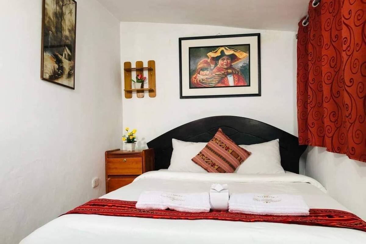 A neatly made bed with white linens and a red patterned pillow, a wooden nightstand with a small vase of flowers, and a framed picture on the wall.