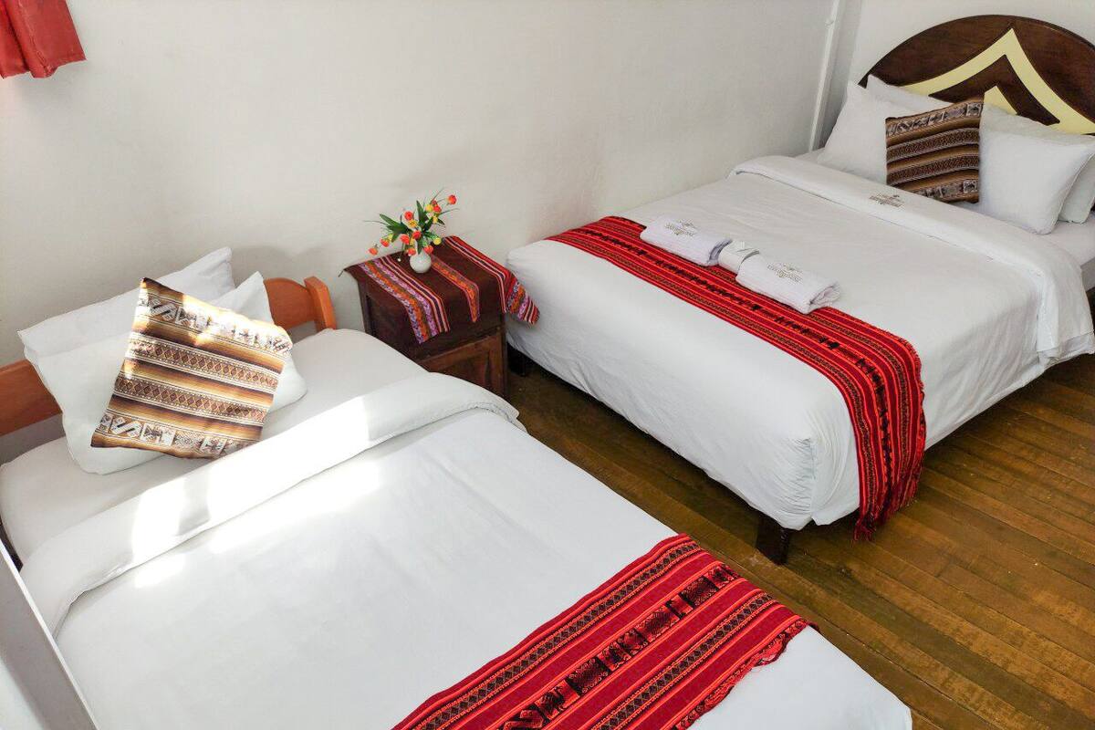 Two single beds with white linens and red decorative borders, a small wooden nightstand with a vase of flowers, and a wooden floor.