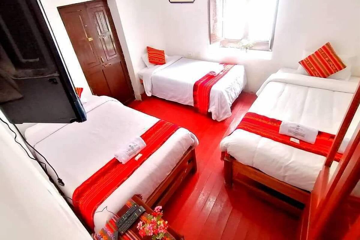 A hotel room with three beds, each with white bedding and red accents. There is a wooden door and a window with a view.