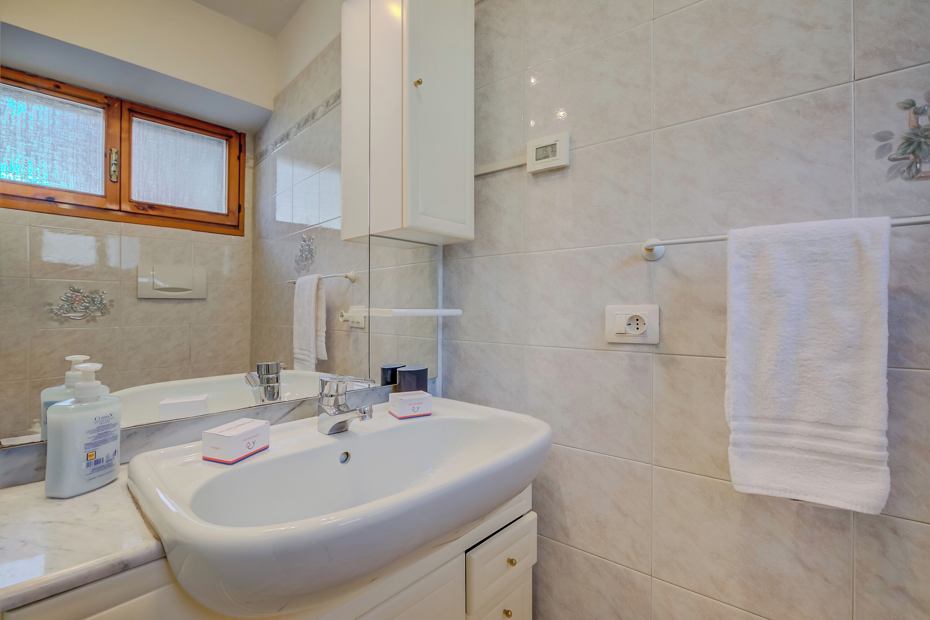 Une salle de bain avec un lavabo blanc, un miroir, une fenêtre et une serviette accrochée au mur.
