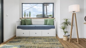 A room with a built-in bench with storage drawers, a large window with a view of palm trees, a standing floor lamp, and a patterned rug.