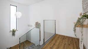 A modern interior with a wooden floor, a glass railing staircase, a hanging light fixture, and a window with a plant.
