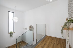 A modern interior with a wooden floor, a glass railing staircase, a hanging light fixture, and a window with a plant.