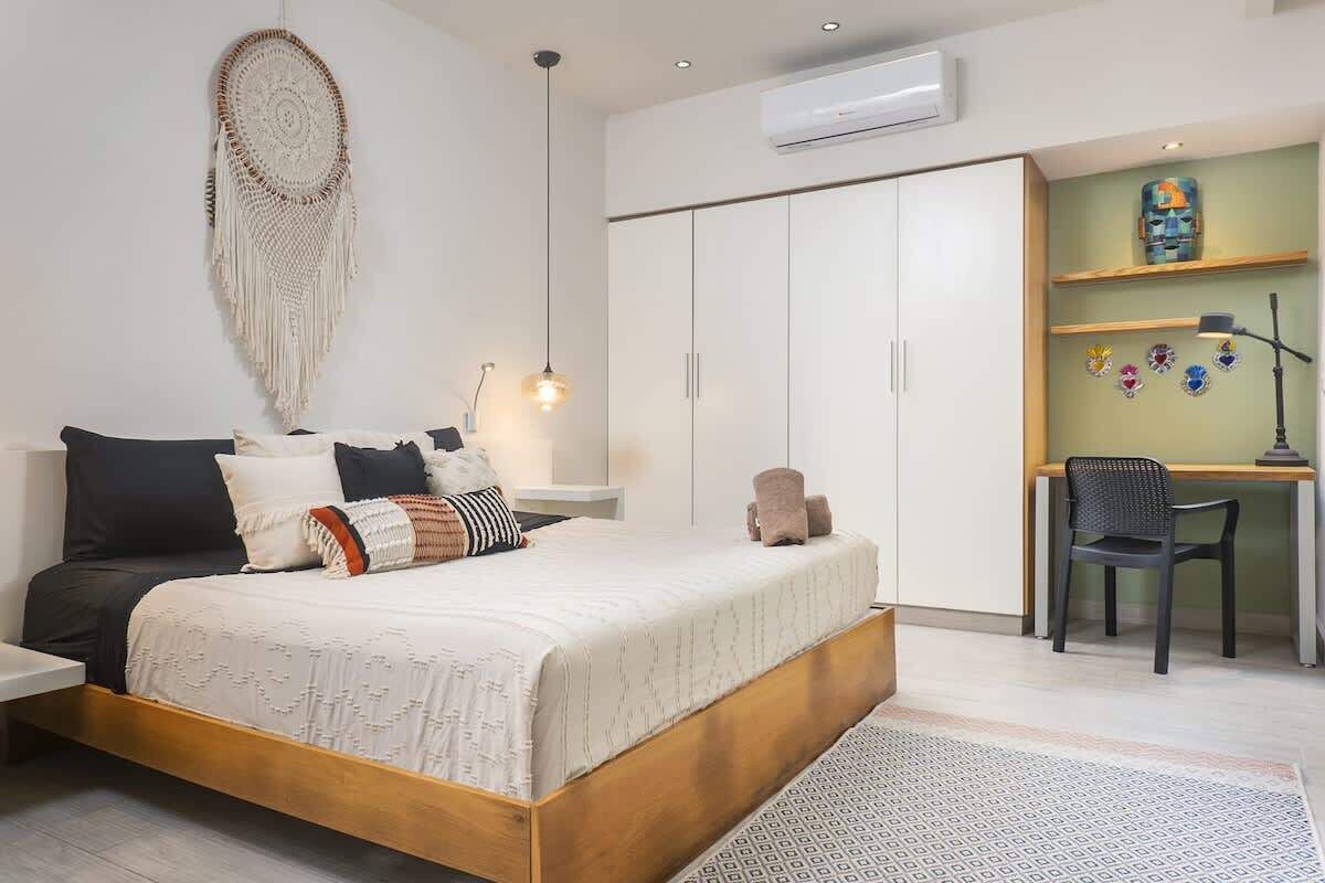 A bedroom with a bed, a desk, and a chair. There is a wall hanging and an air conditioner.