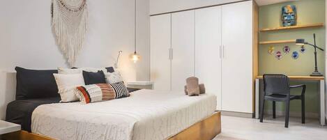 A bedroom with a bed, a desk, and a chair. There is a wall hanging and an air conditioner.