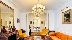 A spacious living room with a fireplace, a chandelier, a sofa, and a dining area in the background.