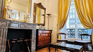 A room with a fireplace, a large ornate mirror, and a chandelier. There are yellow curtains and a view of the outside through a window.