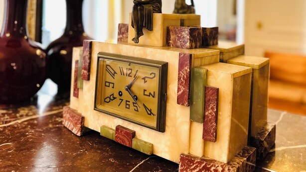 A clock with a Roman numeral display, set on a marble-topped table with decorative blocks.