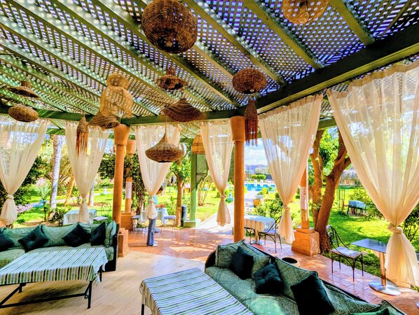 A covered outdoor seating area with wicker hanging lamps, white drapes, and a view of a garden. - Riad Las Palmeras Guest House, Marrakech (Marrakech)