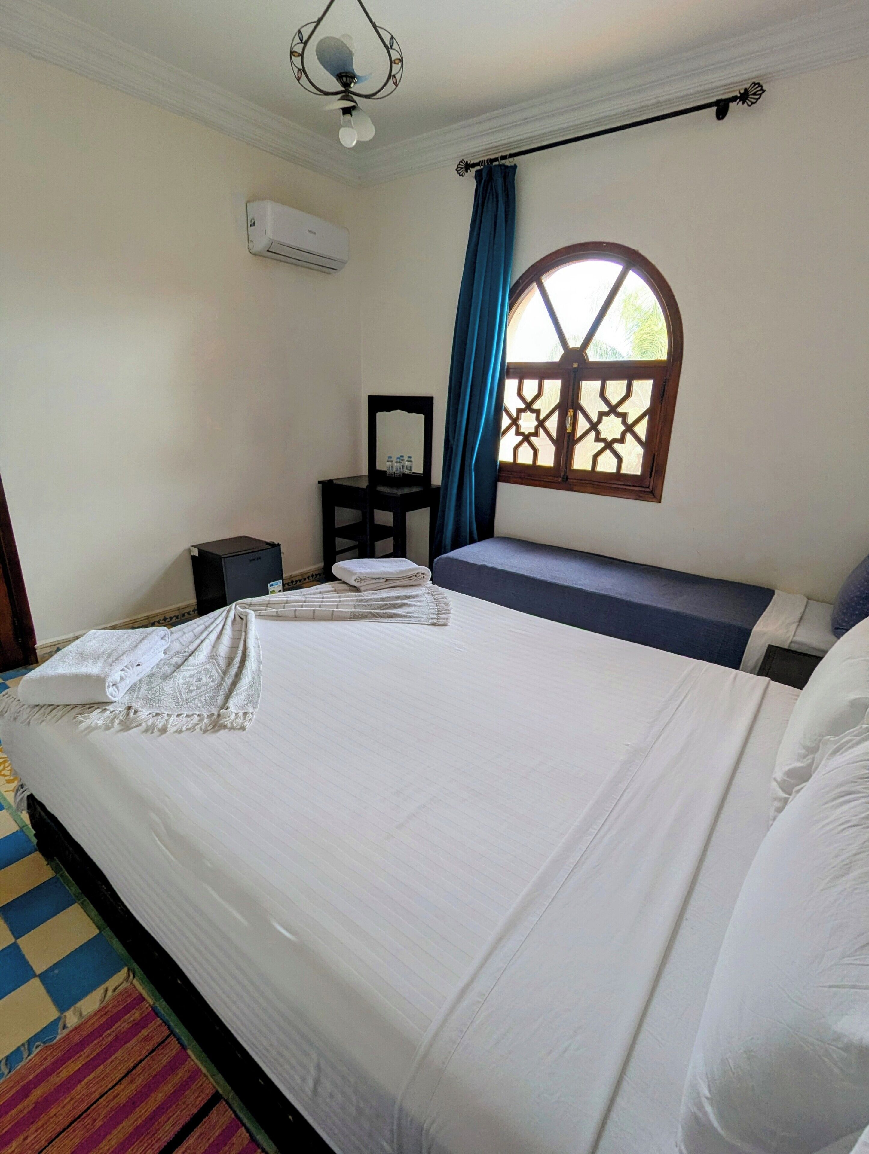 Une chambre avec un grand lit, une petite table avec un miroir, une fenêtre ornée d’un motif géométrique et un appareil de climatisation fixé au mur.