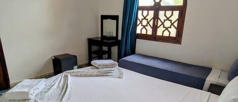 Une chambre avec un grand lit, une petite table avec un miroir, une fenêtre ornée d’un motif géométrique et un appareil de climatisation fixé au mur.