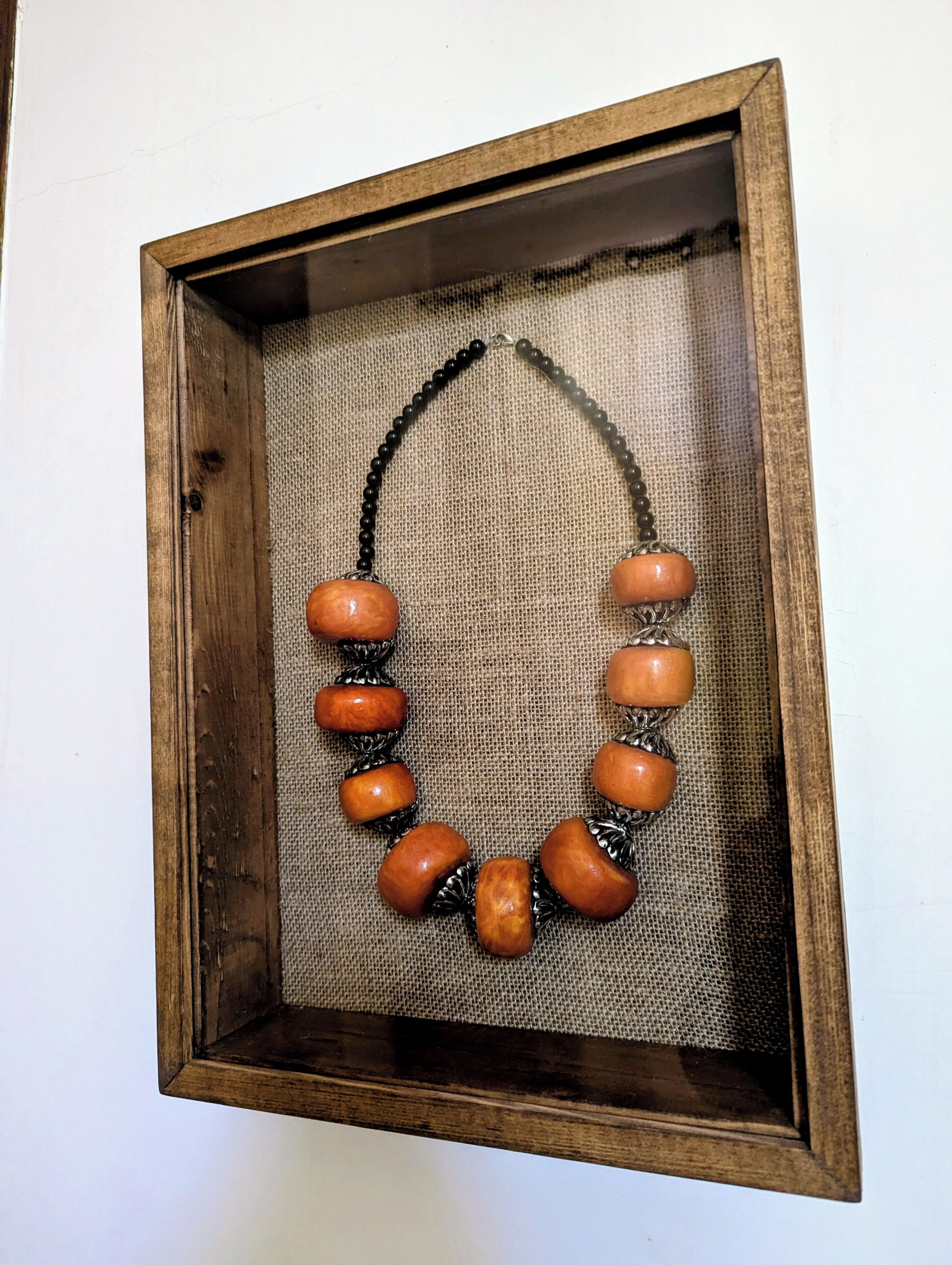 A necklace with wooden beads and black accents, displayed in a wooden frame.