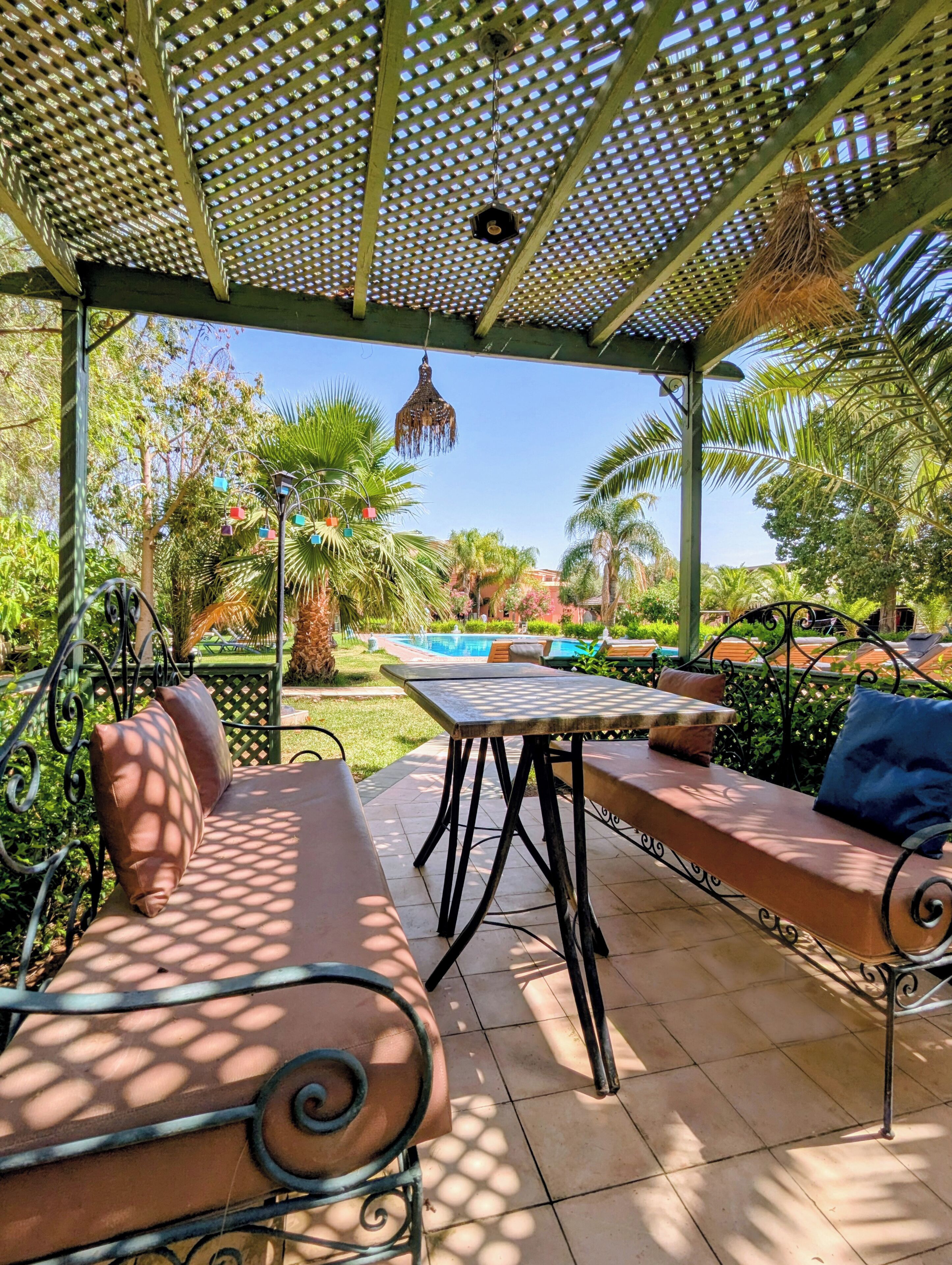 An outdoor patio area with a pergola, seating, and a view of a pool and greenery.