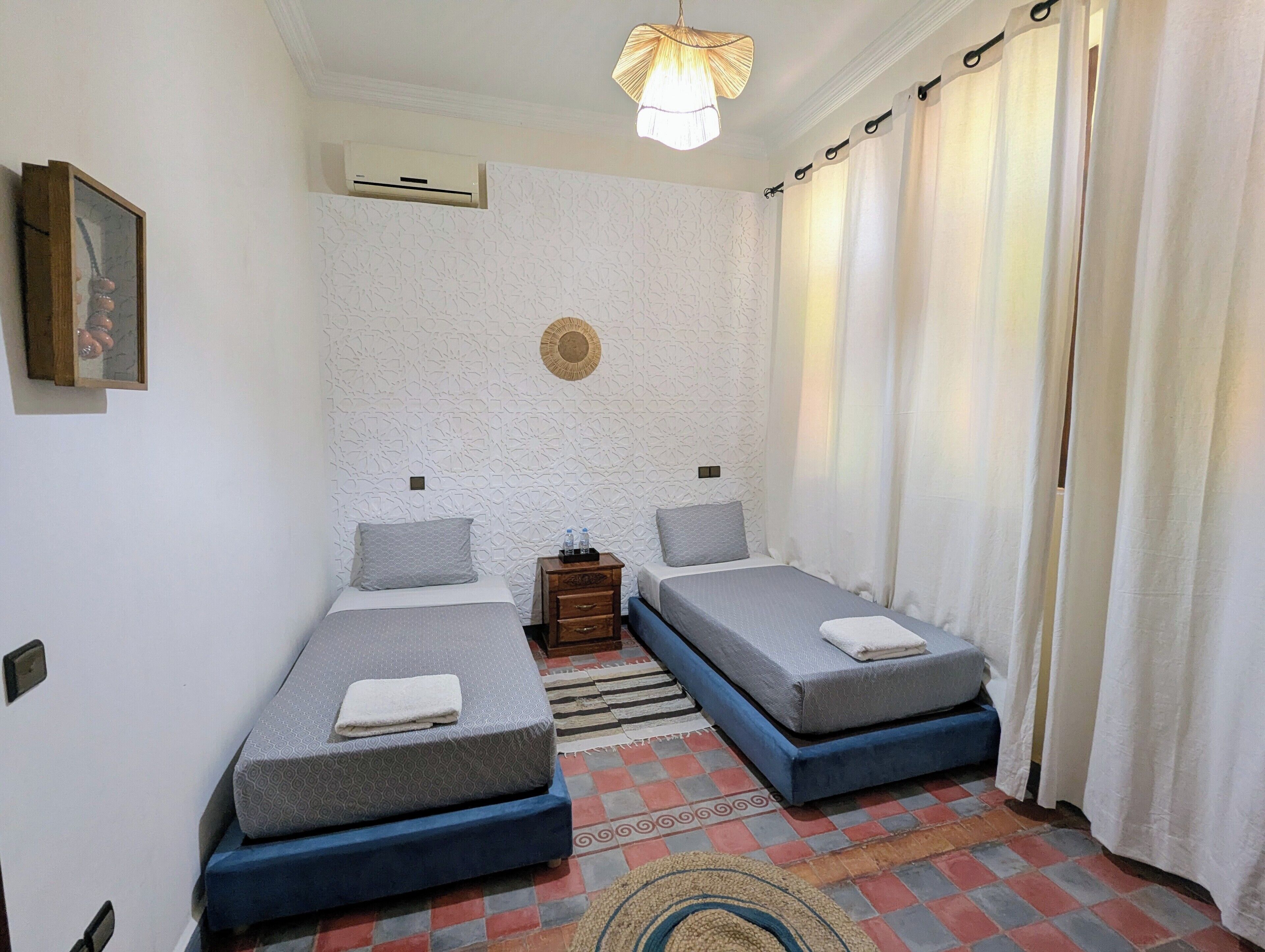 A room with two beds, a small wooden nightstand, a wall-mounted air conditioner, a patterned rug, and a ceiling light fixture.