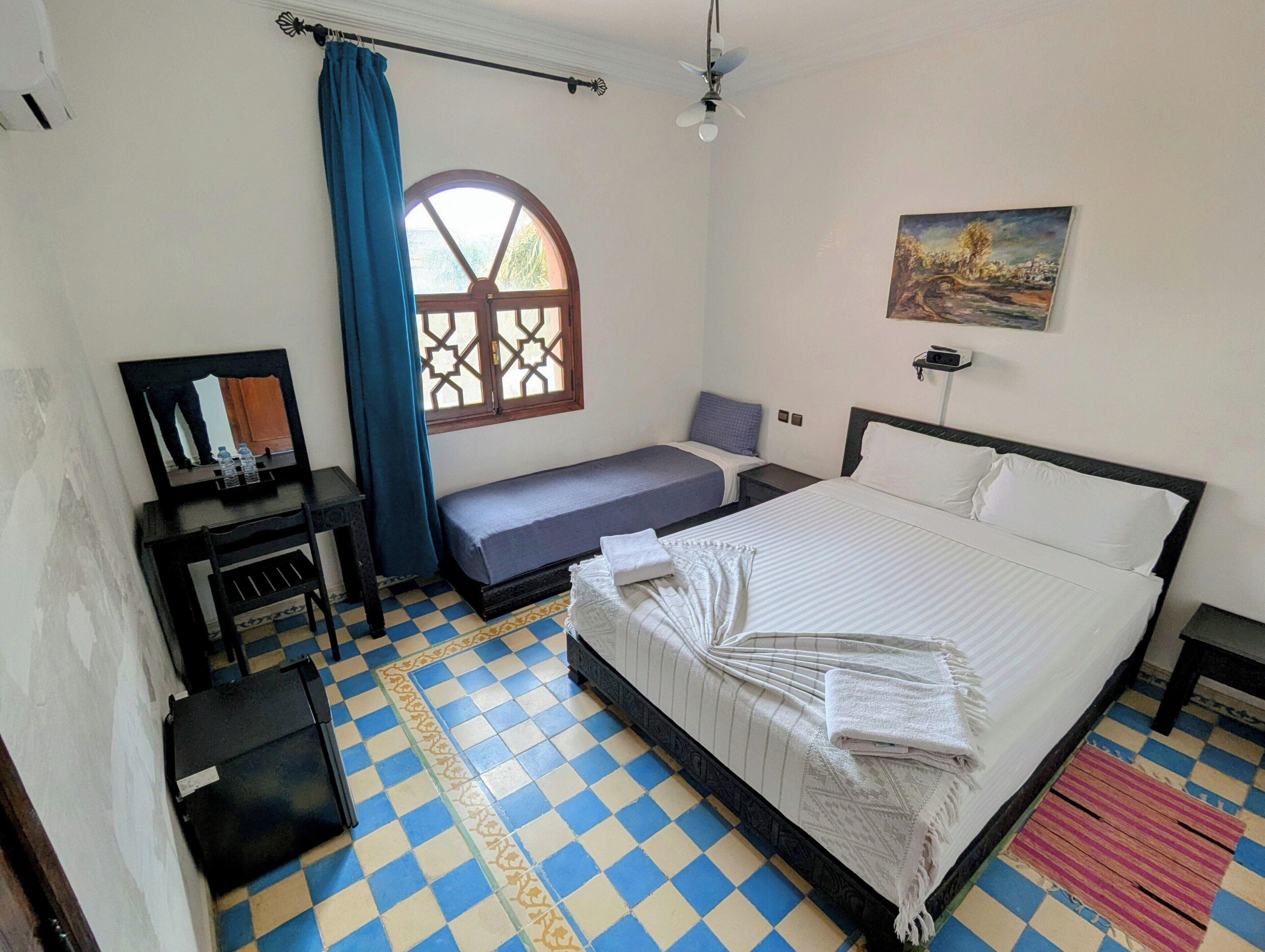 A room with a checkered floor, a bed, a bench, a desk, a window with a curtain, and a painting on the wall.