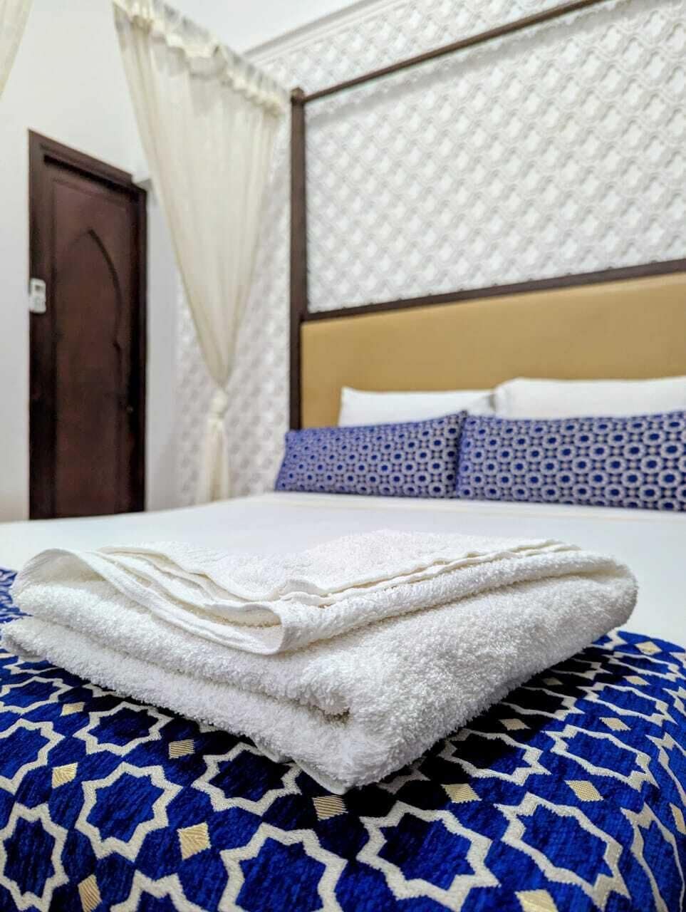 Una cama bien hecha con una toalla blanca doblada, una colcha de diseño azul y negro, y un cabecero con textura.