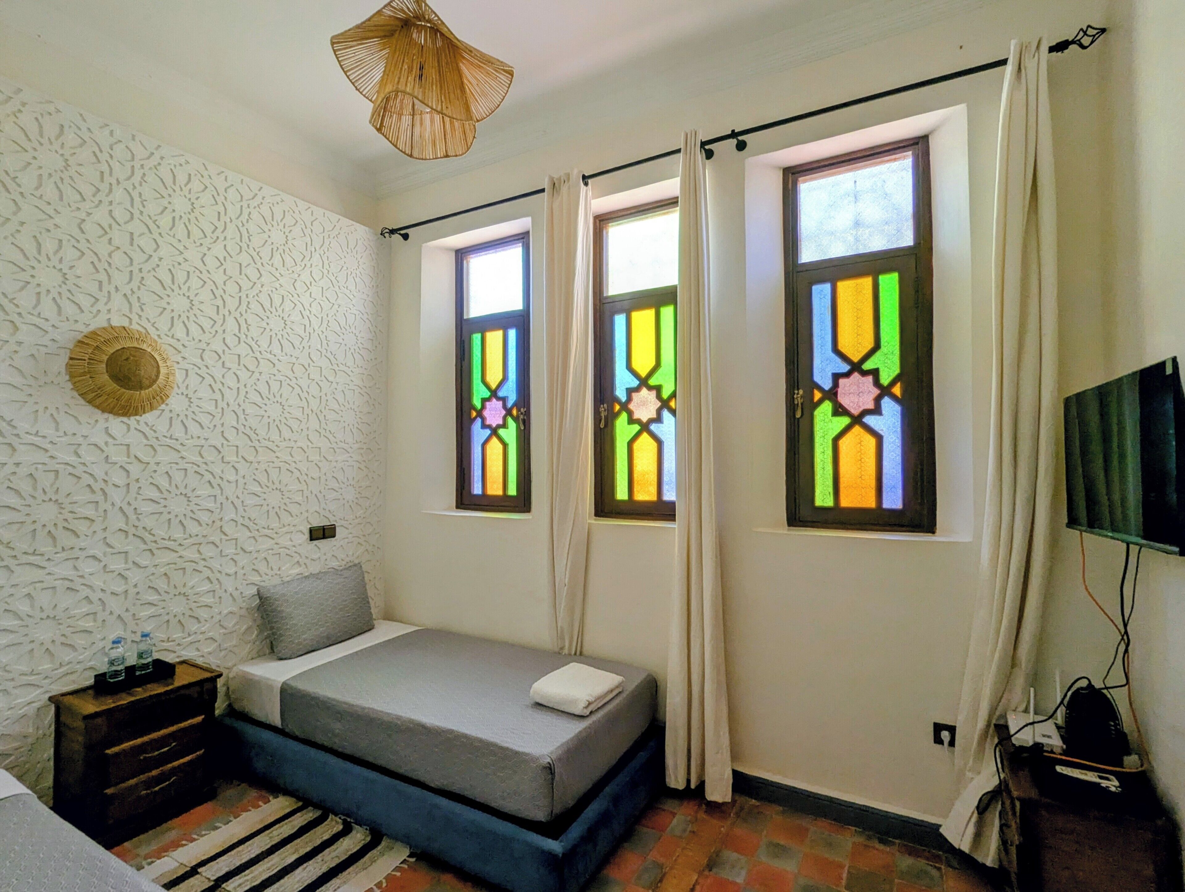 A bedroom with a bed, a nightstand, a TV, and two windows with stained glass.