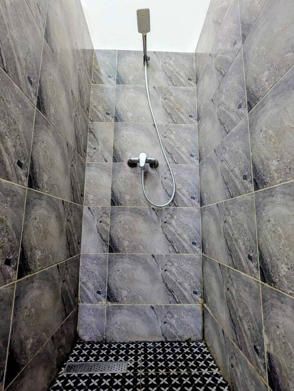 Eine Dusche mit Handbrause und wandmontiertem Wasserhahn, in einem Raum mit Marmoroptik an den Wänden und einem gemusterten Boden.