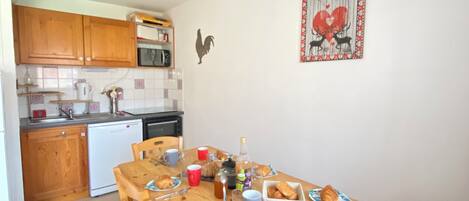 A compact kitchen with wooden cabinets, a microwave, and a sink. A dining table is set with plates, cups, and a bottle.