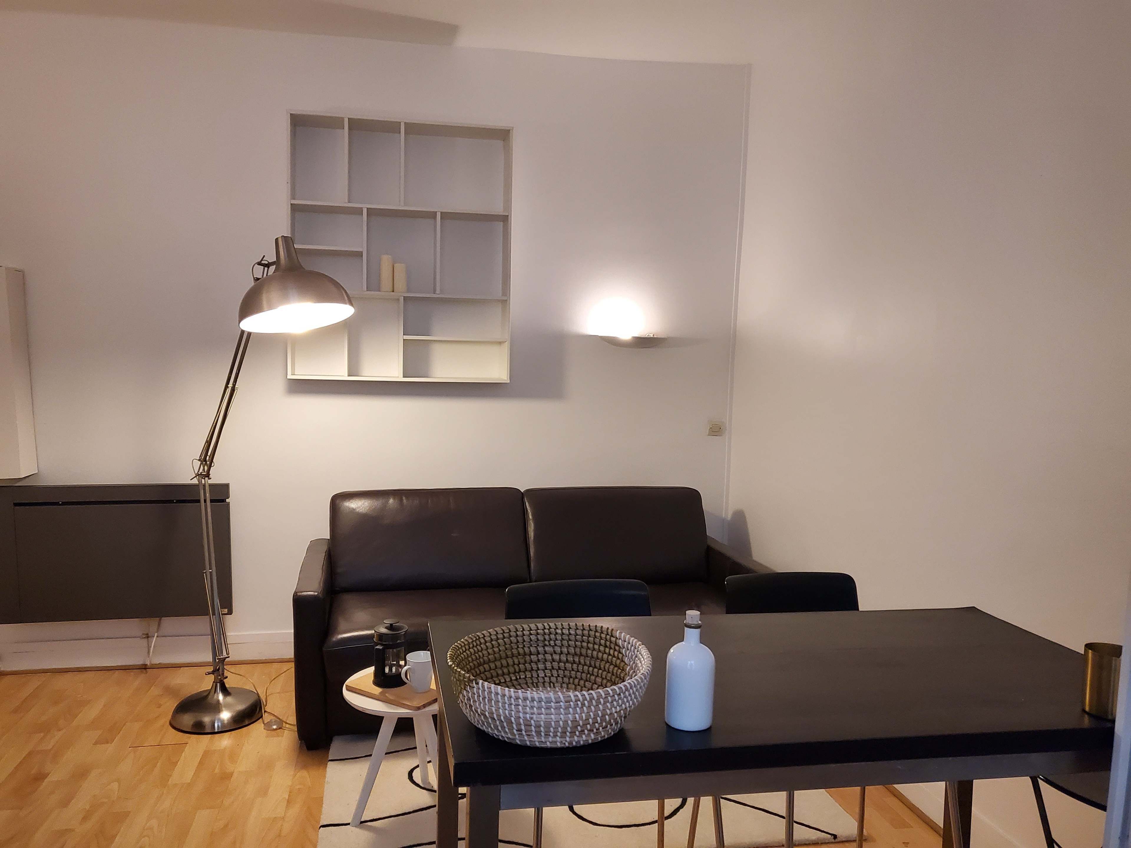 Un salon moderne avec un canapé en cuir noir, une table à manger surmontée d’un panier en osier, une lampe sur pied et une étagère fixée au mur.