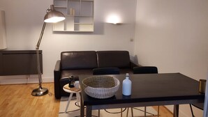 A modern living room with a black leather sofa, a dining table with a woven basket, a floor lamp, and a wall-mounted shelf.