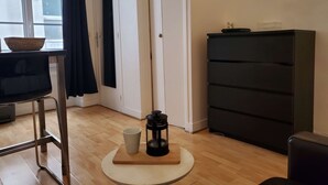 A room with a wooden floor, a round table with a coffee maker and a cup, a black chair, a black cabinet, and a window with a view of buildings.