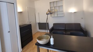 A small, modern living room with a black sofa, a wooden table, a black cabinet, and a white wall-mounted shelf.
