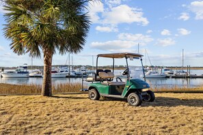 A golf cart parked on a sandy area with a body of water and boats in the background.