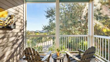 A covered outdoor seating area with Adirondack chairs and a small table, overlooking a residential street and greenery.