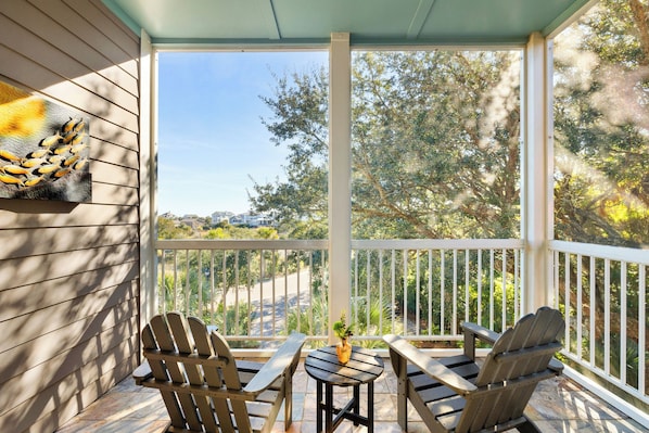 A covered outdoor seating area with Adirondack chairs and a small table, overlooking a residential street and greenery. - Regatta Inn | The Catalina - Relaxing B&B on Folly Island (Folly Beach)