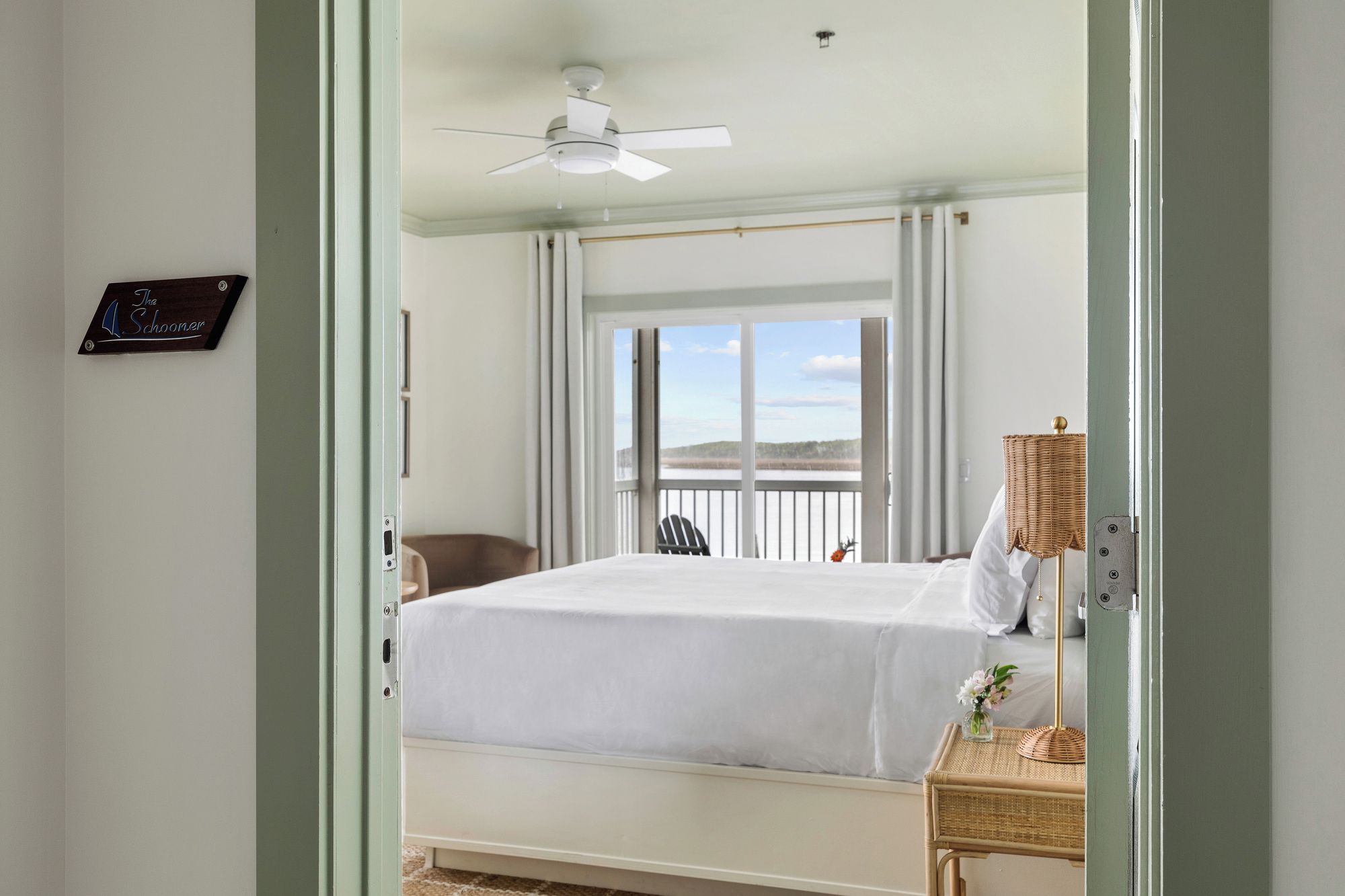 A bedroom with a bed, a ceiling fan, a window with a view, and a wicker side table with a lamp.