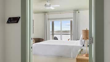A bedroom with a bed, a ceiling fan, a window with a view, and a wicker side table with a lamp.