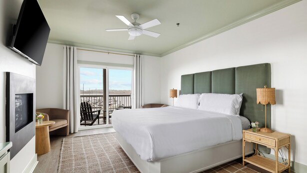 Habitación de hotel con cama, televisor, ventilador de techo, mesita de noche, lámpara y vista al exterior.