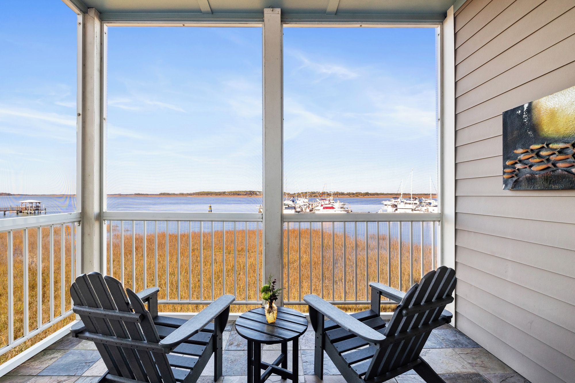 A covered outdoor seating area with two Adirondack chairs and a small table, overlooking a marina with boats and a distant shoreline.