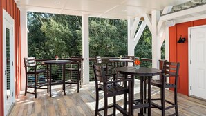 Un patio al aire libre con mesas y sillas de madera, rodeado por una estructura blanca con puertas de cristal.