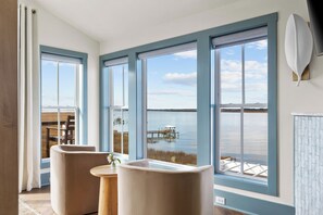 Ein Zimmer mit großen Fenstern und Blick auf ein Gewässer, ein Holztisch mit einer Vase sowie zwei Stühle.